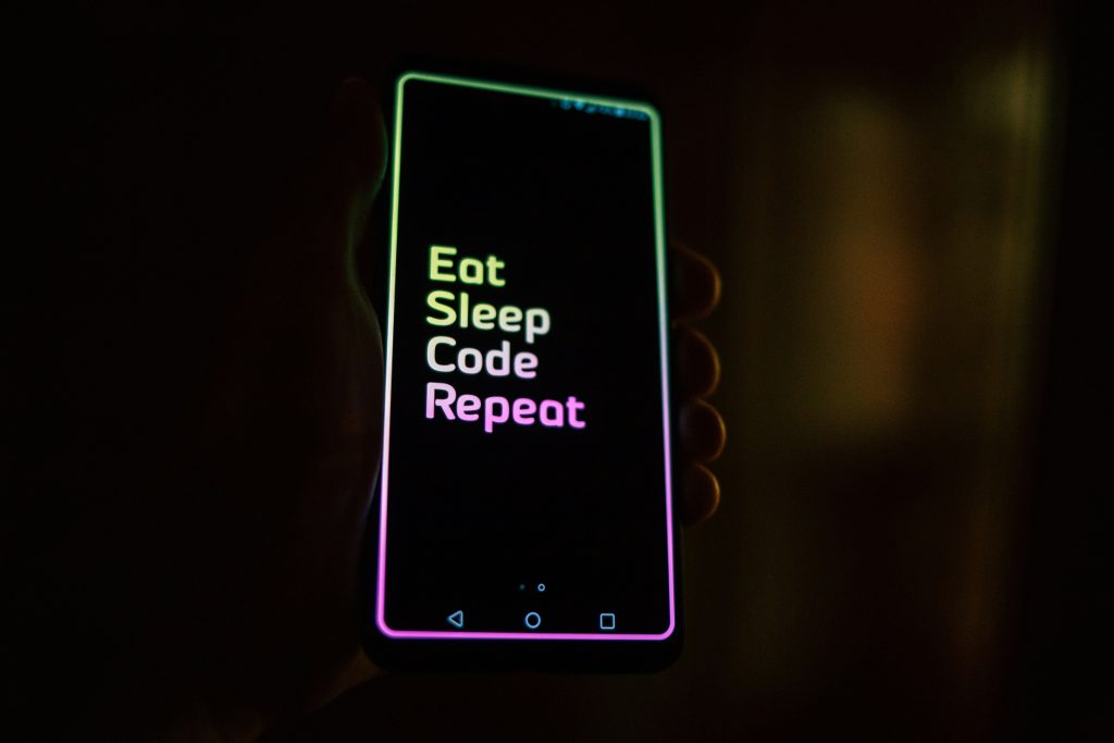A phone in darkness turned on and held by a hand that says "Eat Sleep Code Repeat"
